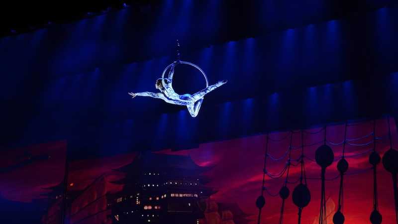 Acrobat high in the air, holding a hoop, in front of a virtual set.